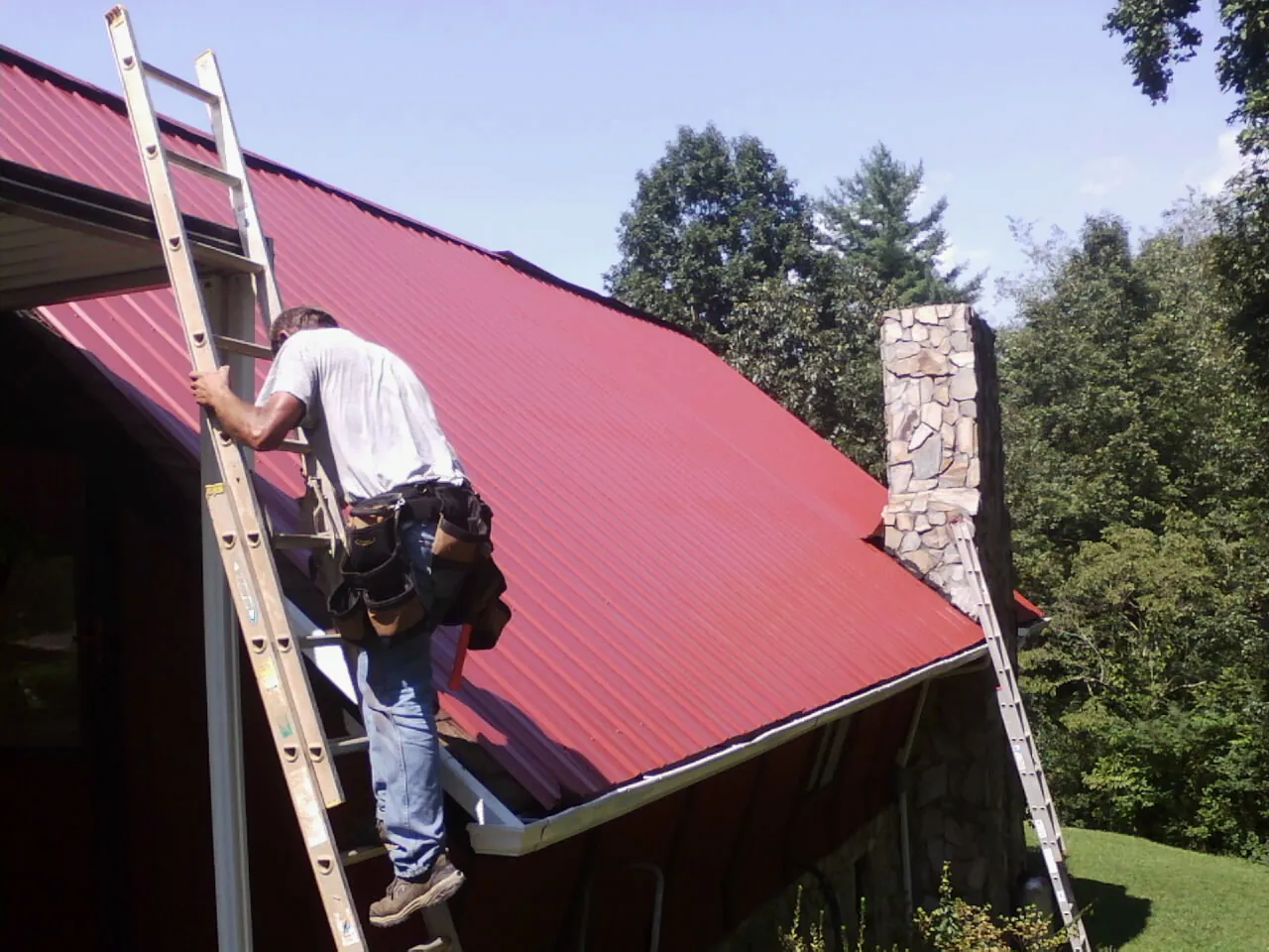 Roofer on a ladder beside a red metal roof and stone chimney — Soffit & Fascia Repair by Roofing Brothers in South Rockwood